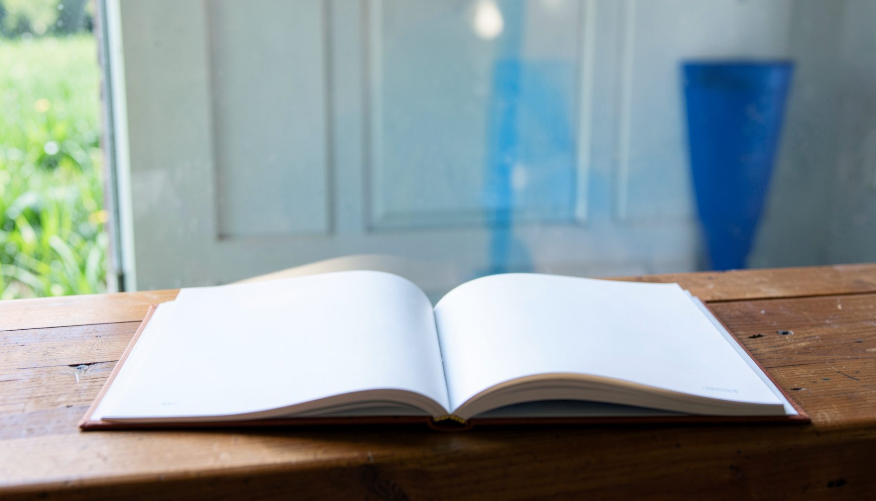 Отворена книга на дрвена маса покрај прозорец со поглед кон зелени полиња, осветлена со мека дневна светлина во природни тонови